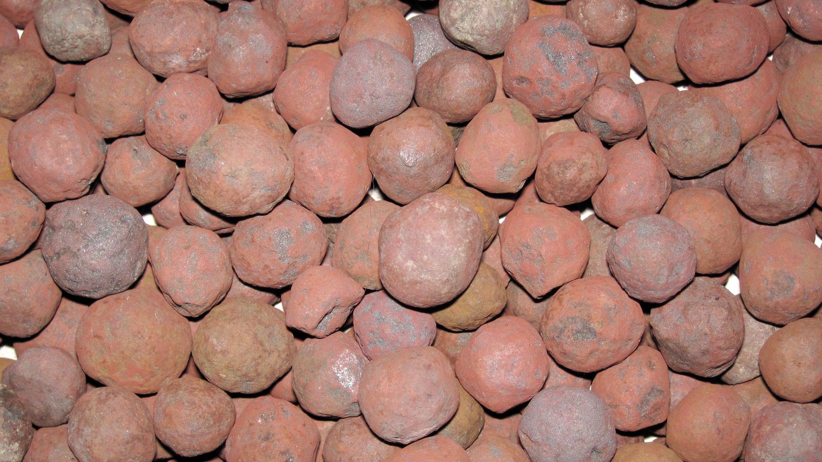 A dense mass of rust red iron ore pellets, roughly marble sized and pitted, packed together and filling the whole frame.