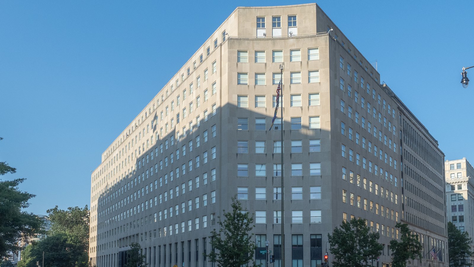 The limestone Export-Import Bank Building on Vermont Avenue in Washington, seen from the street corner under a clear sky, with an American flag on a pole at the corner and street trees along the pavement.