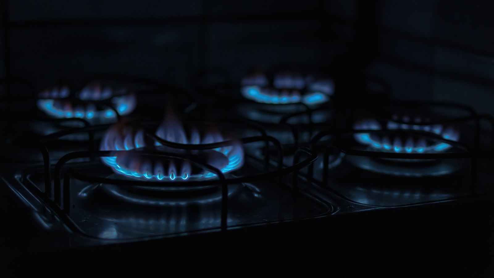 Four gas burner rings alight in a darkened room, low blue flames tipped with orange under black pan supports, the rest of the frame in shadow.