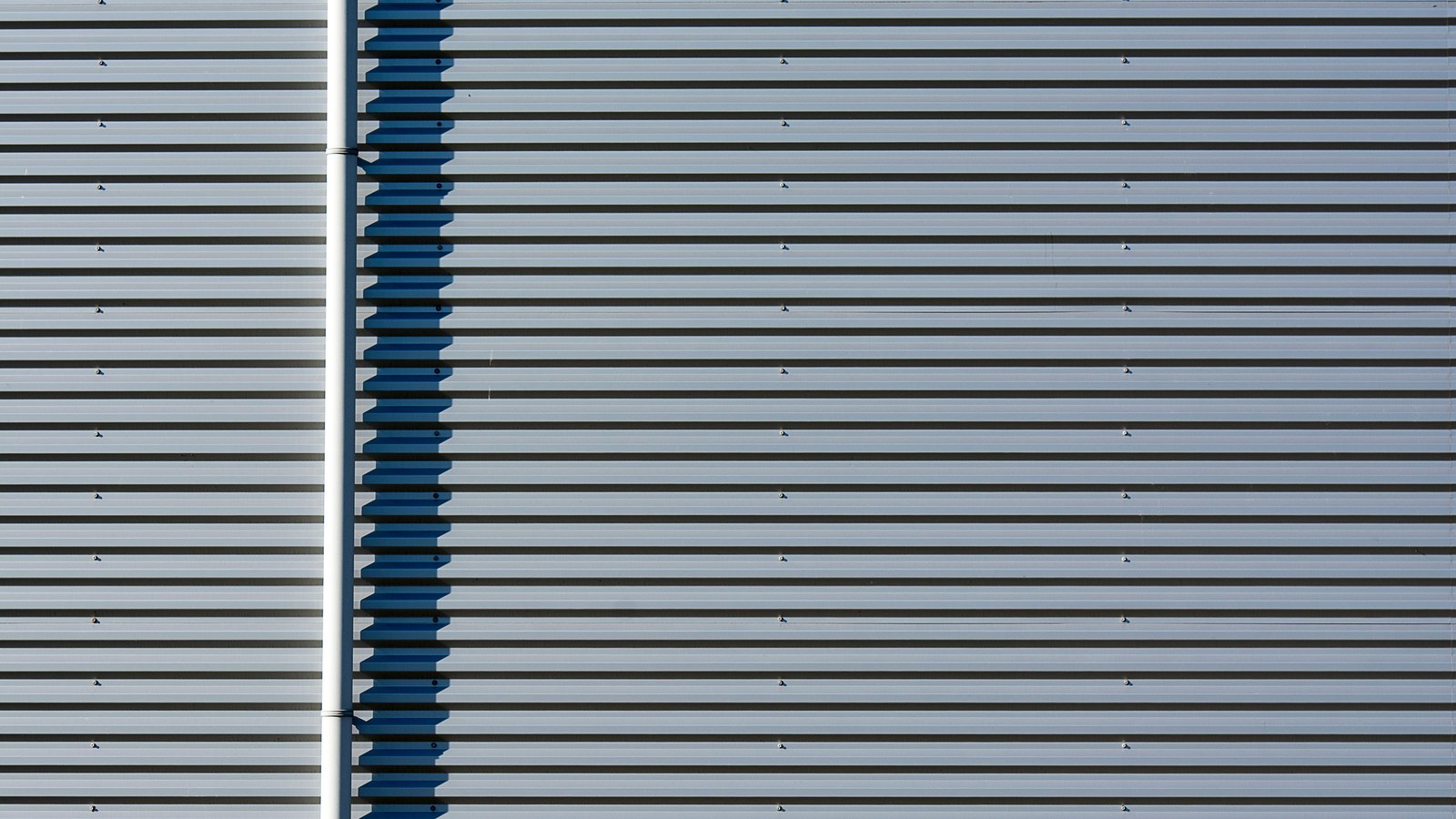 A close view of a ribbed metal building wall, pale horizontal cladding panels crossed by a narrow blue vertical strip and a white downpipe.
