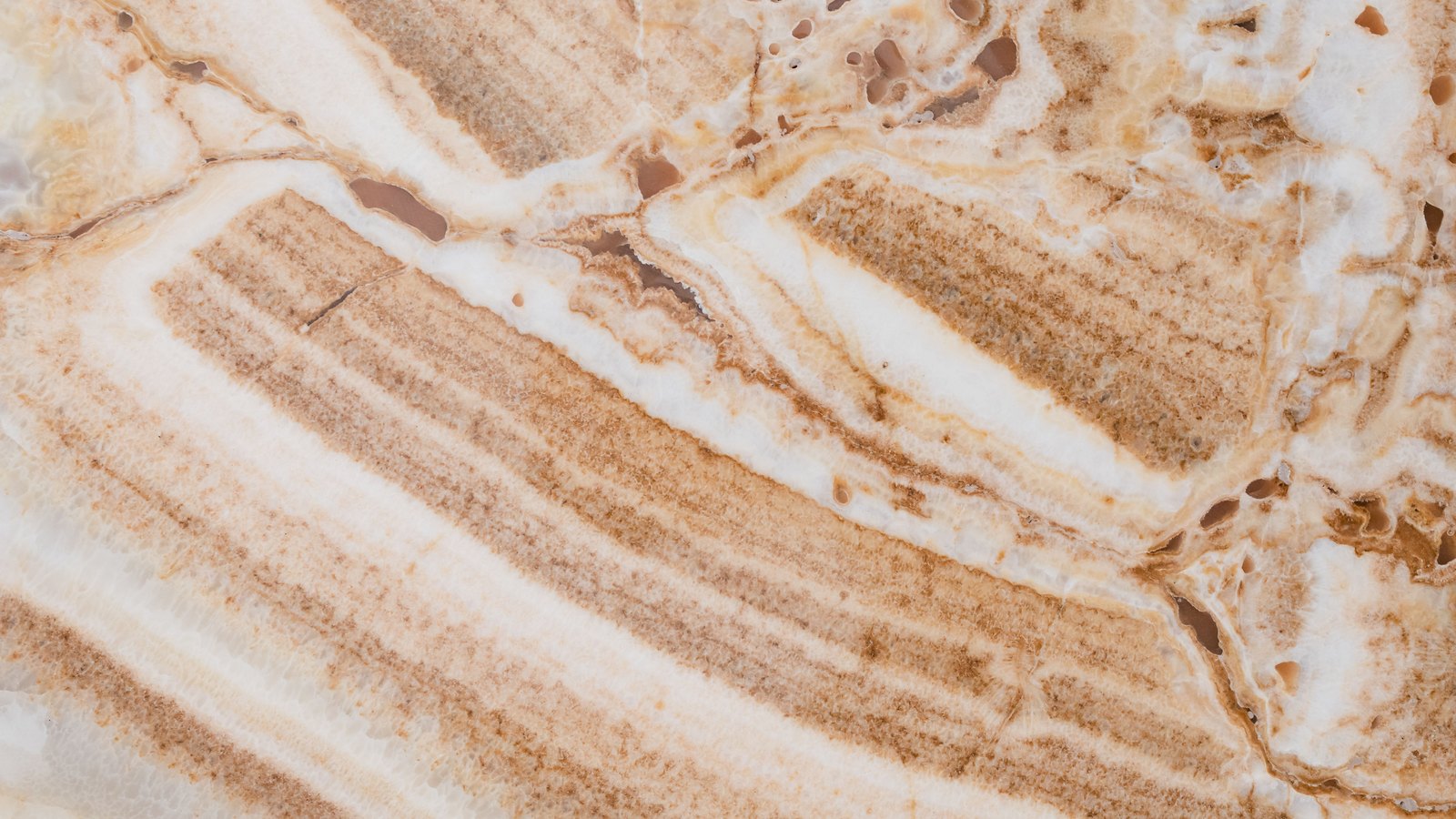 A close photograph of a polished slab of banded cream and white onyx, the layers running diagonally across the frame with rust-brown mineral bands between them and small dark cavities in the stone towards the upper right.