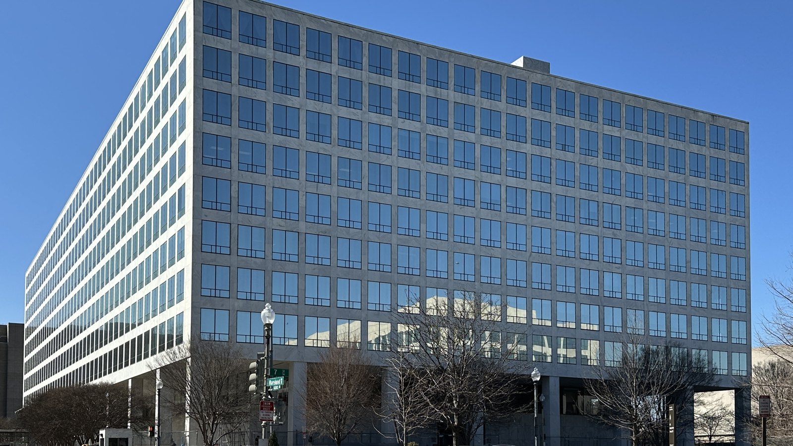 The Orville Wright Federal Building in Washington, which houses the Federal Aviation Administration, seen from a street corner: a nine storey slab clad in pale stone with a regular grid of dark tinted windows, lifted clear of the ground on piers, with bare winter trees, street lamps and a traffic signal along the pavement in front of it under a clear blue sky.