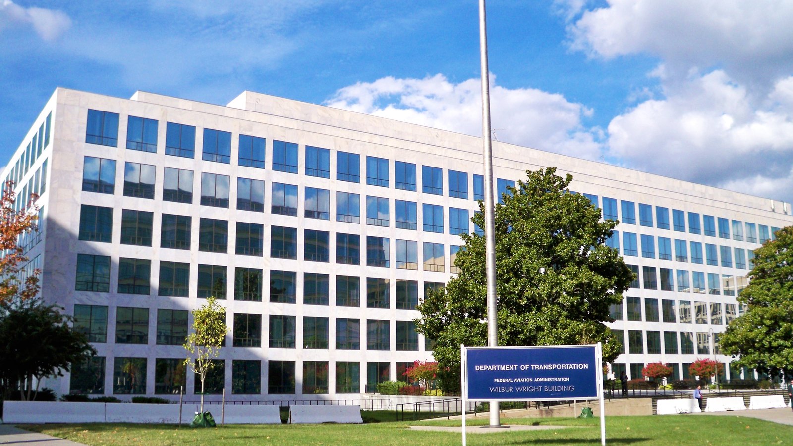 A long white stone office building of six storeys with dark windows, seen from a lawn behind trees under a blue sky with scattered cloud. A flagpole stands in front of it and a blue sign on the grass reads Department of Transportation, Federal Aviation Administration, Wilbur Wright Building. Two people stand at a distance near the entrance.