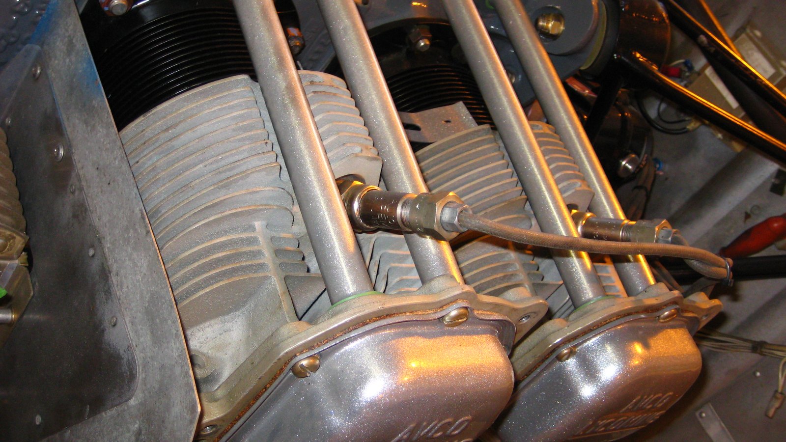 A close view inside an opened engine cowling at two finned aluminium cylinders lying on their sides, with push rod tubes running diagonally across them and two polished rocker covers in the foreground carrying the cast maker's name.