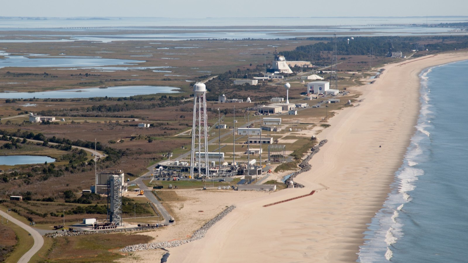 An aerial photograph of the launch pads and support buildings at Wallops Island on the Virginia coast, hemmed between marsh on one side and a narrow strip of beach and the Atlantic on the other.