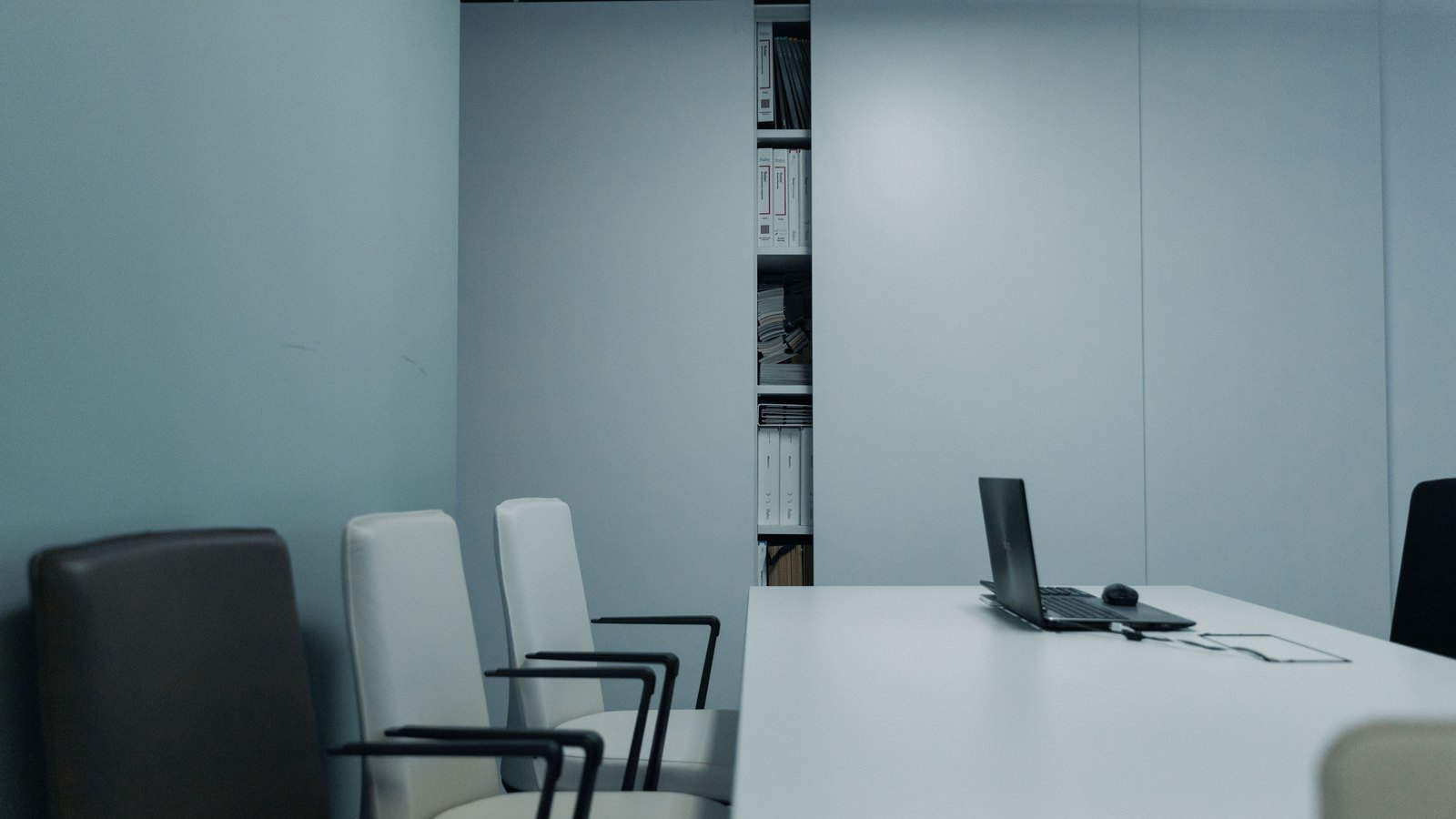 An empty conference room with white chairs around a long table and an open laptop at the far end.