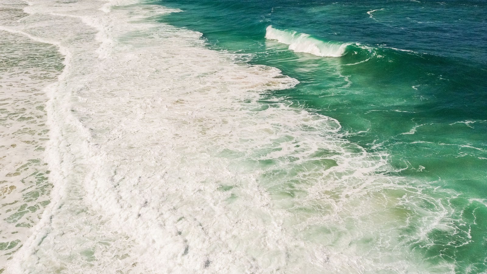 An aerial view looking straight down at ocean surf breaking in white lines across turquoise and green water beside a pale sandy shore, with no people, boats, aircraft, buildings, signage, text or identifiable landmark anywhere in the frame. A generic illustration of the water alongside a beach, not a photograph of this event.