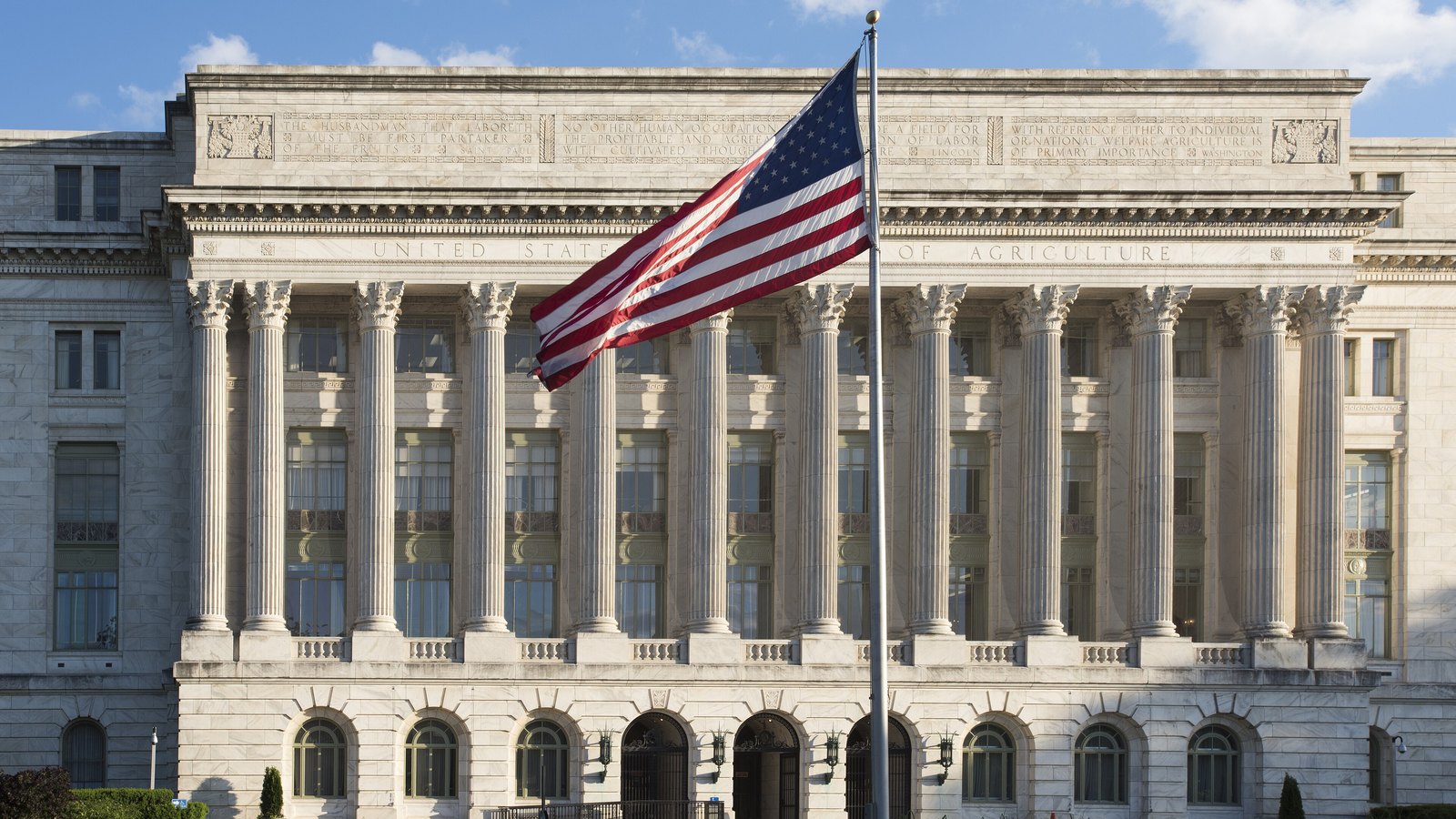 The Jamie L. Whitten Building, headquarters of the United States Department of Agriculture in Washington: a white marble facade behind a long row of Corinthian columns, the words United States Department of Agriculture carved across the entablature, quotations cut into the storey above, and an American flag flying from a pole in front of it.