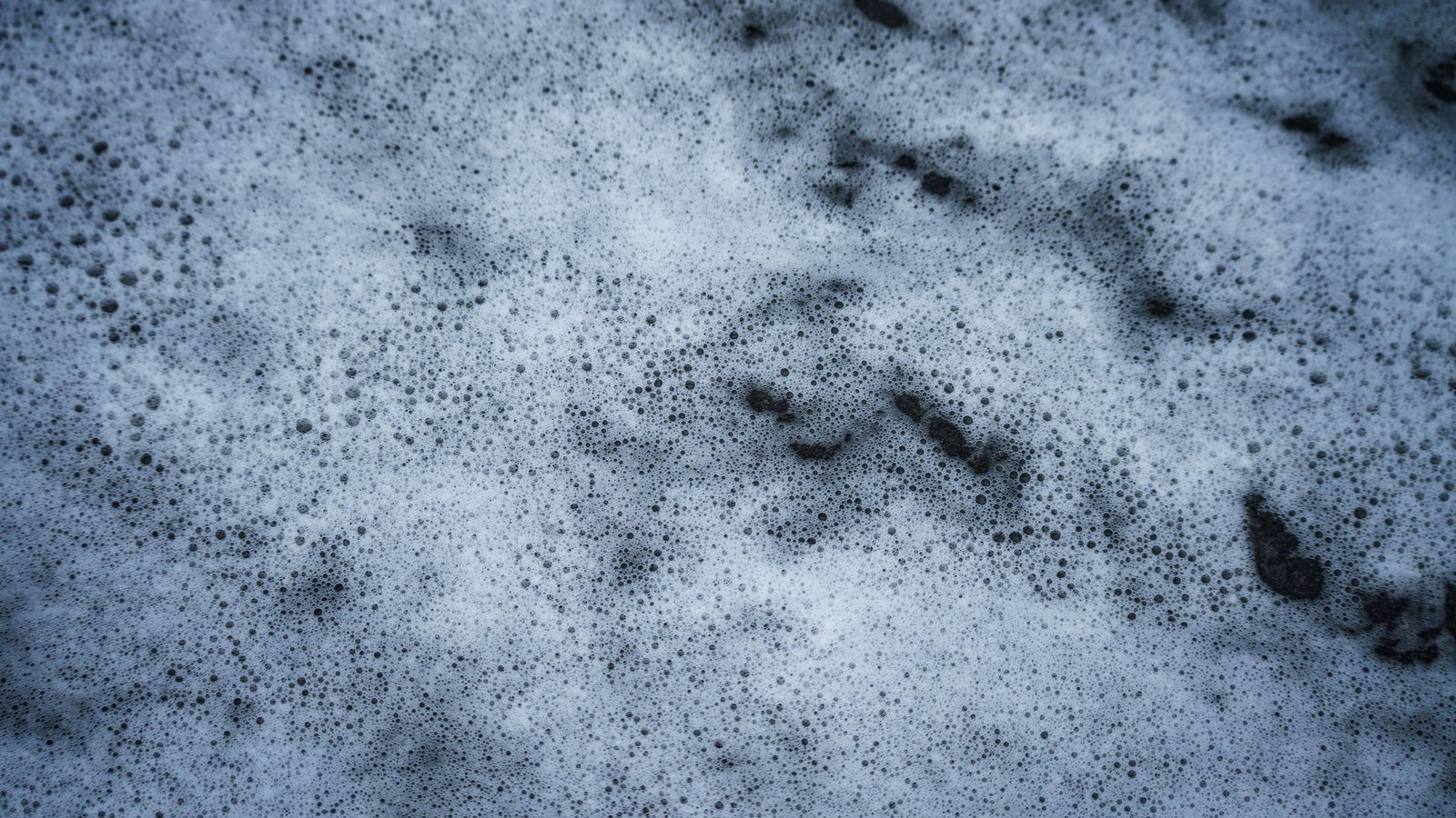 A close overhead view of white soap foam on a dark wet surface, filling the frame: a dense mat of fine bubbles, thickest and palest through the middle, thinning towards the top right where the dark surface shows through.