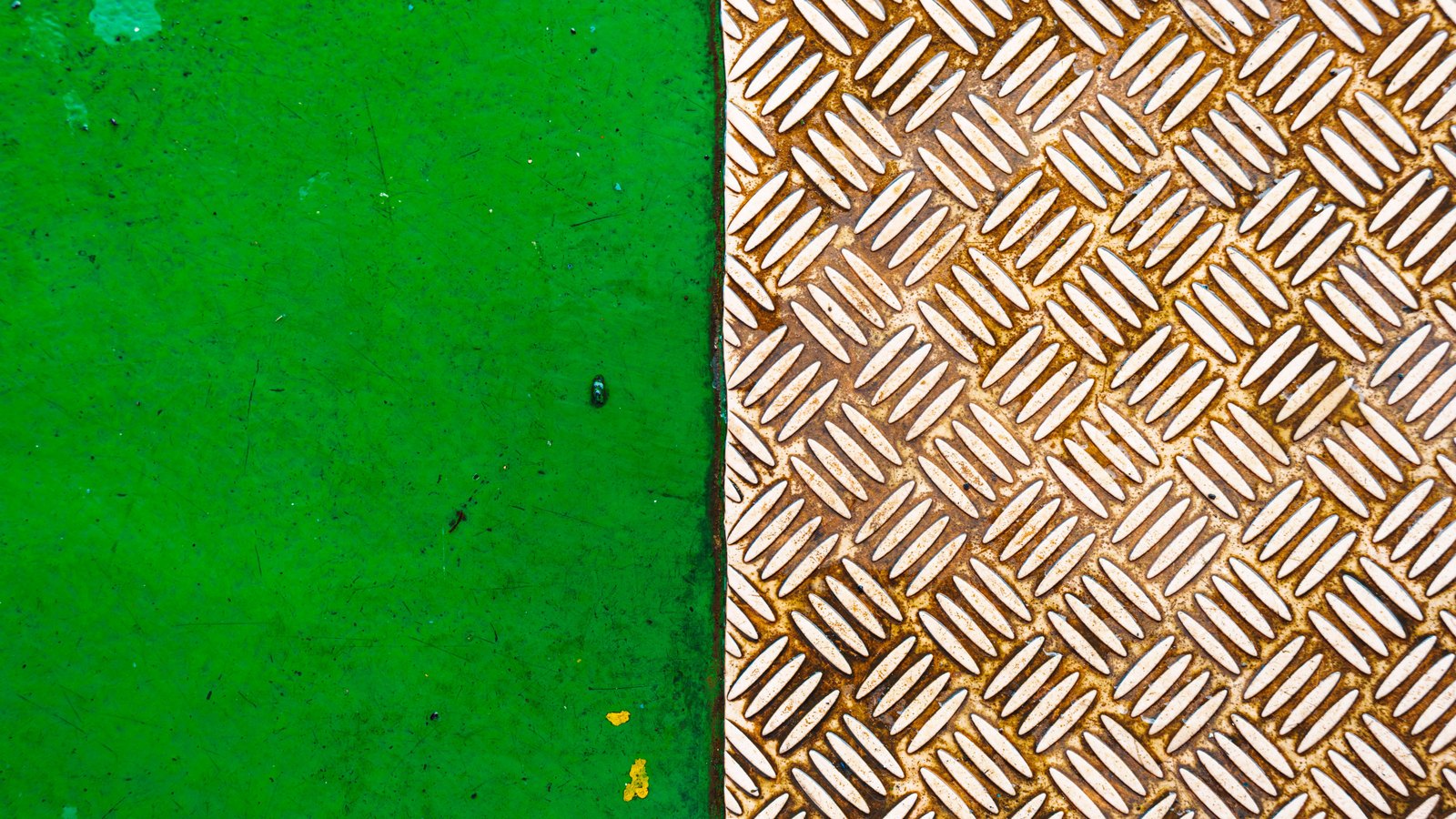 A close overhead photograph of an industrial steel floor plate, the right half raised in a repeating diamond tread pattern with orange corrosion spread through the channels between the studs, the left half a flat panel finished in worn green paint and meeting the tread along a straight seam. No object, person, place, marking or lettering is in view.