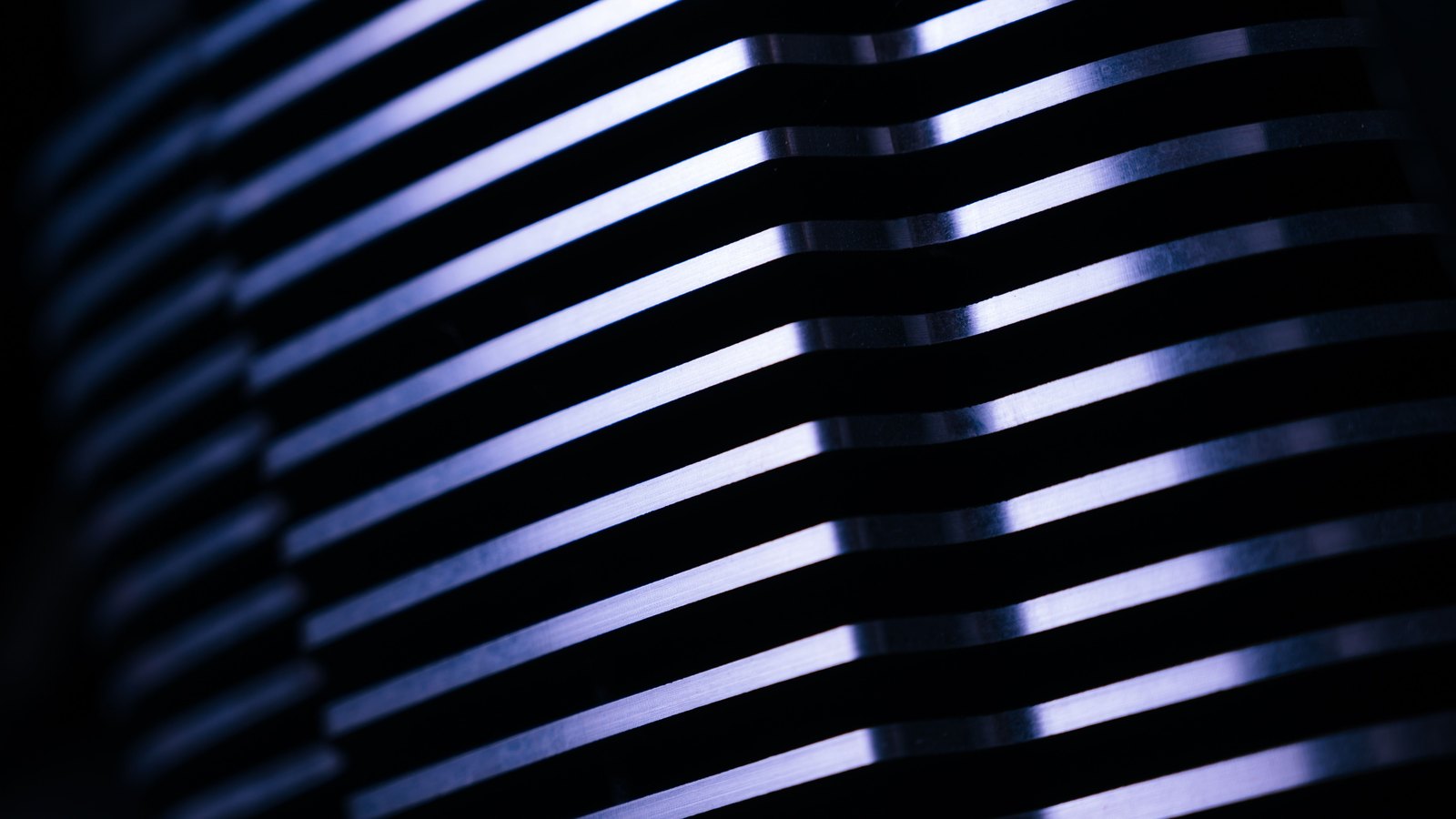 A tight photograph of the stacked fins of an aluminium heat sink curving away into darkness, each machined edge catching a cold blue and white highlight, with no lettering, brand or hand in the picture.