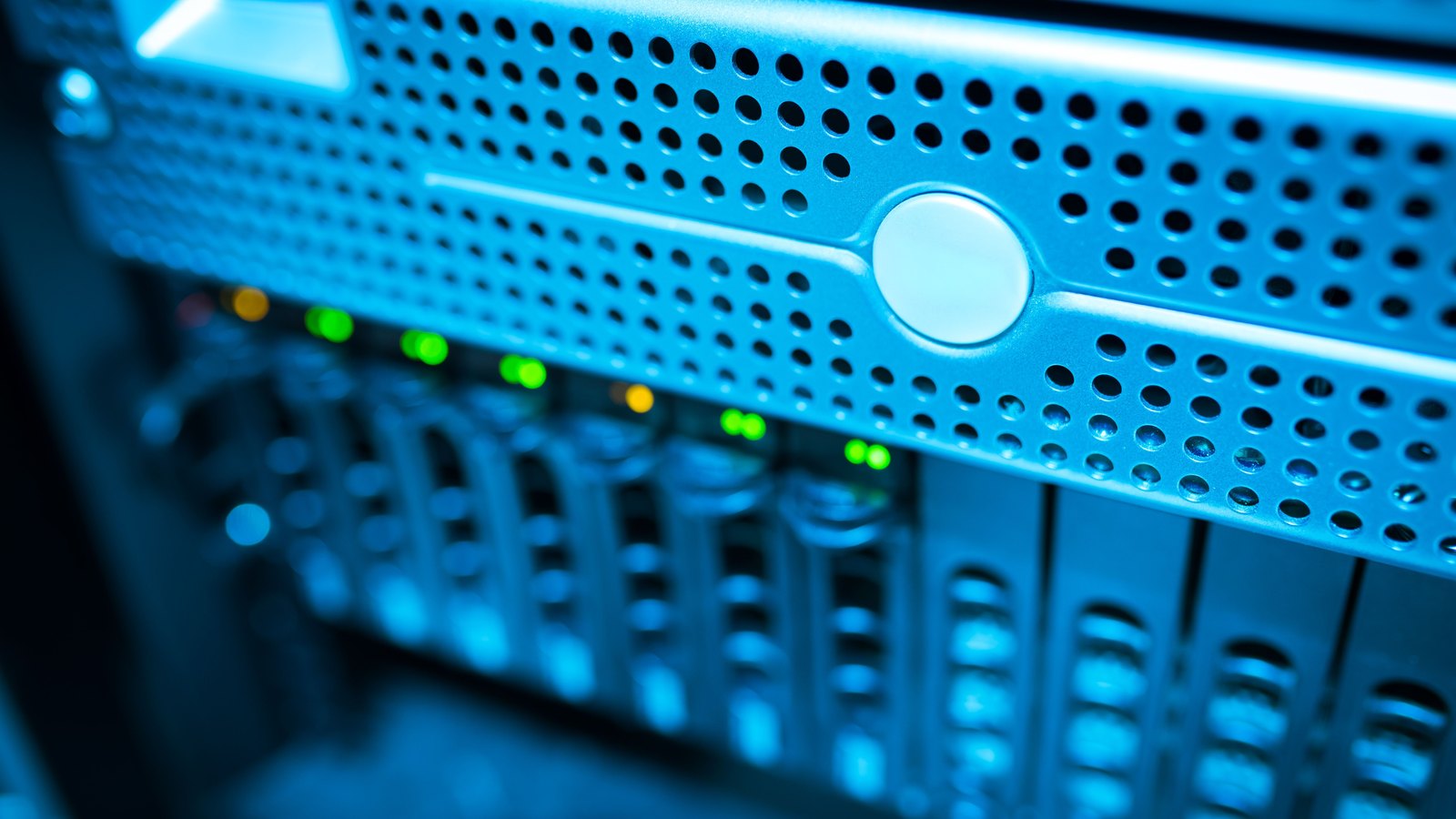 A tight close-up of the front of a rack mounted server lit in blue, its perforated metal face filling the upper half of the frame and a row of small green and amber status lights running beneath it, with further drive bays blurred behind. No person, screen content, logo, serial number or legible text of any kind appears in the frame. A generic illustration of the carrier network equipment a breach notification rule governs, not a photograph of any particular company's systems.