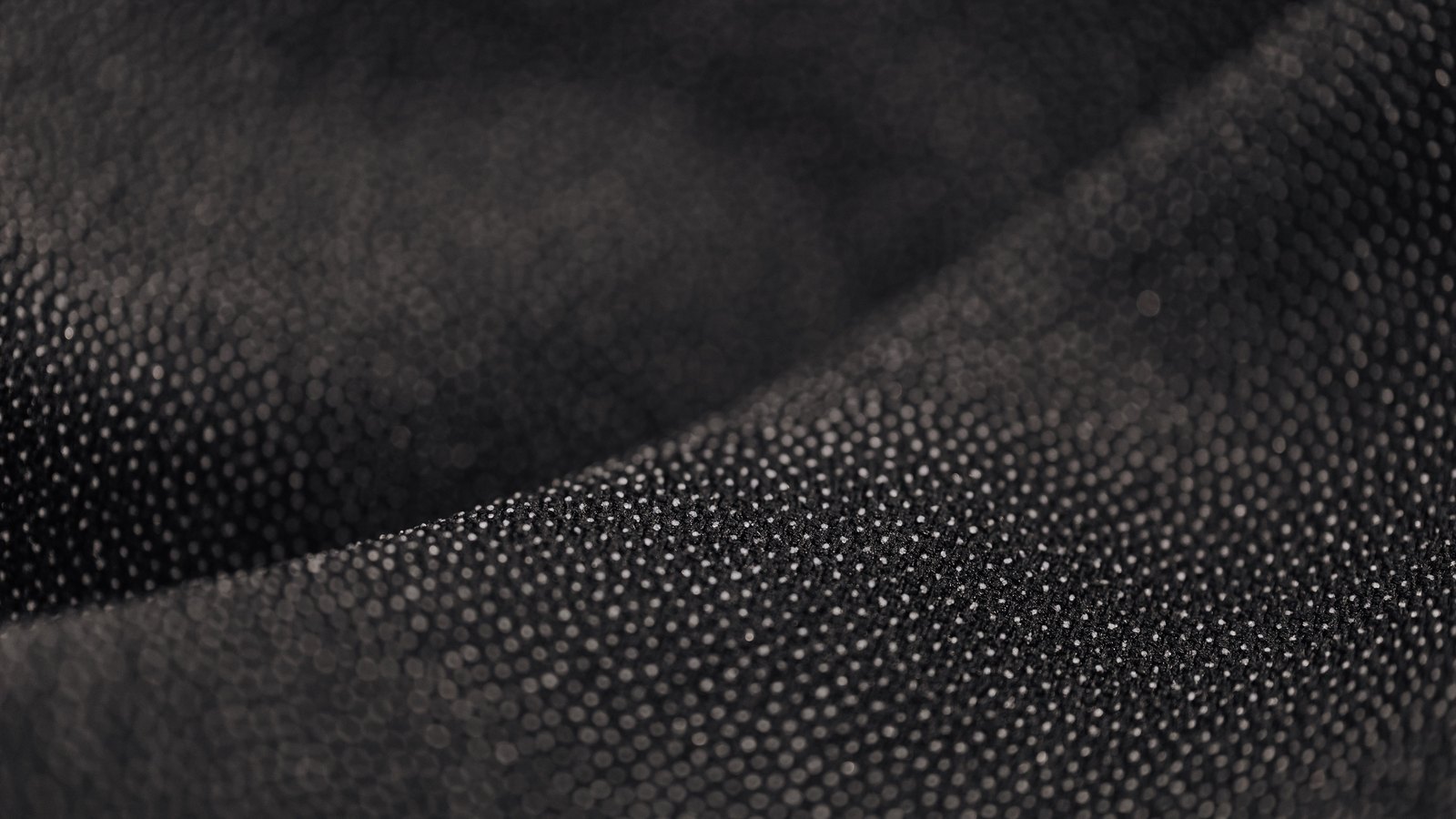 A close photograph of black cloth lying in two soft diagonal folds and filling the frame, the surface densely covered in tiny pale dots that catch the light along the raised fold and fade into shadow at the top left. No object, person, marking, lettering or brand mark is in view.