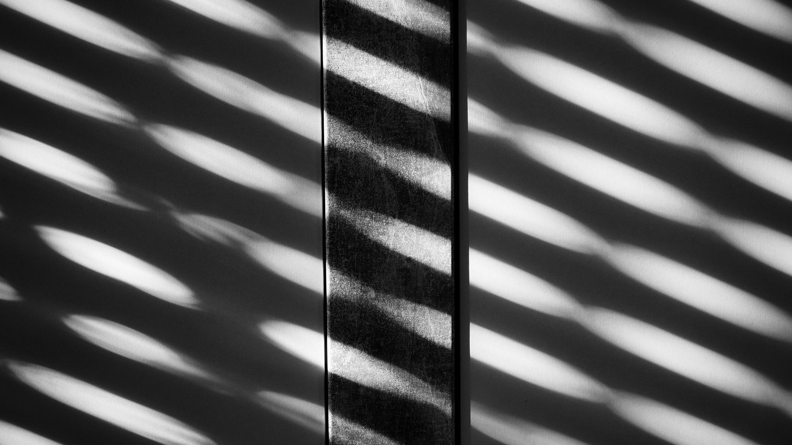 Diagonal bands of light and shadow falling across a plain interior wall in black and white, broken by a single vertical post running down the centre of the frame.