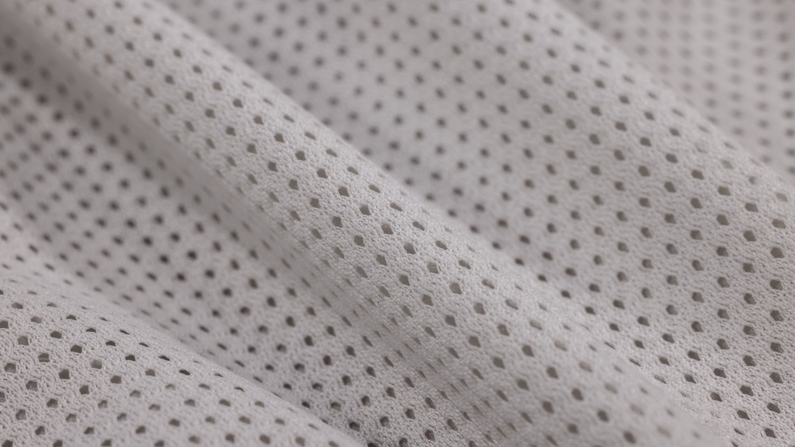 A close photograph of white perforated mesh fabric filling the whole frame, small round holes in even rows running over soft diagonal folds in flat light. No text, brand mark, person or location is visible.