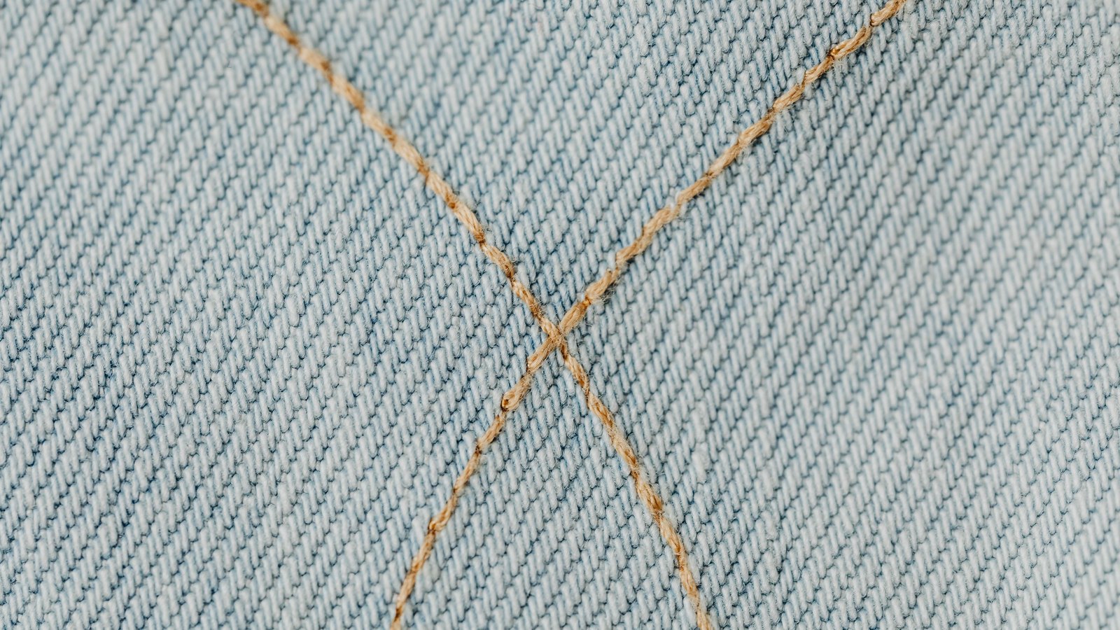 A macro view of pale blue denim, its fine diagonal twill lines running corner to corner, crossed near the centre by two lines of tan topstitching that meet in an X. No text, brand mark, person or place is in view.