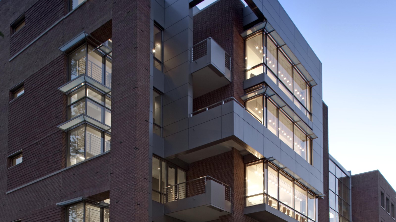 A corner of a Food and Drug Administration building on the White Oak campus in Maryland at dusk: dark red brick and grey metal panelling, projecting glazed bays with lit ceilings visible inside, small railed balconies set between them, and a clear evening sky behind. There is no signage in the frame.