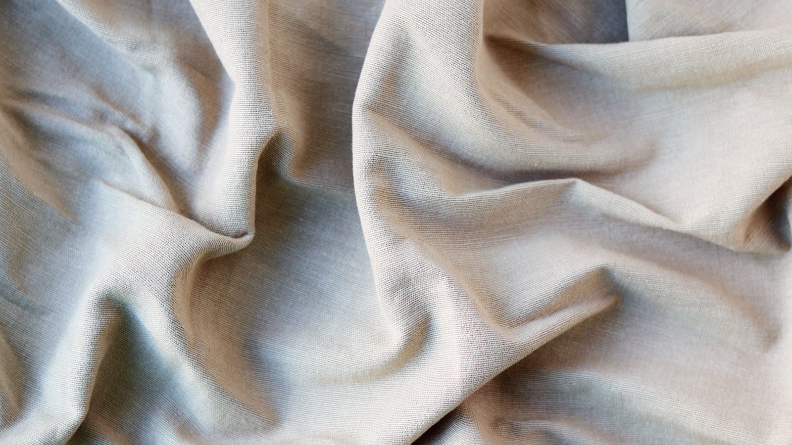 A close photograph of pale cream cotton cloth gathered into soft folds that fill the frame, the weave showing as a fine even grain across the raised surfaces and shadow pooling in the deeper creases toward the left. No object, person, marking, lettering or brand mark is in view.