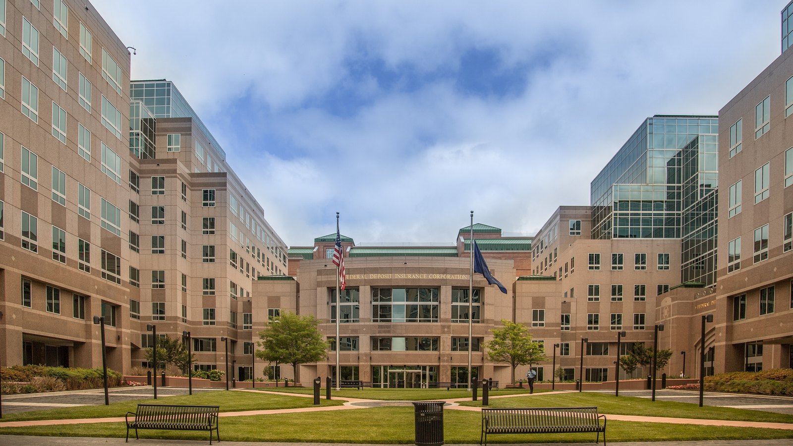A beige stone and glass office campus arranged around a lawn with benches and young trees, the words Federal Deposit Insurance Corporation cut into the stone above the central entrance, with an American flag and a second flag on poles in front of it.