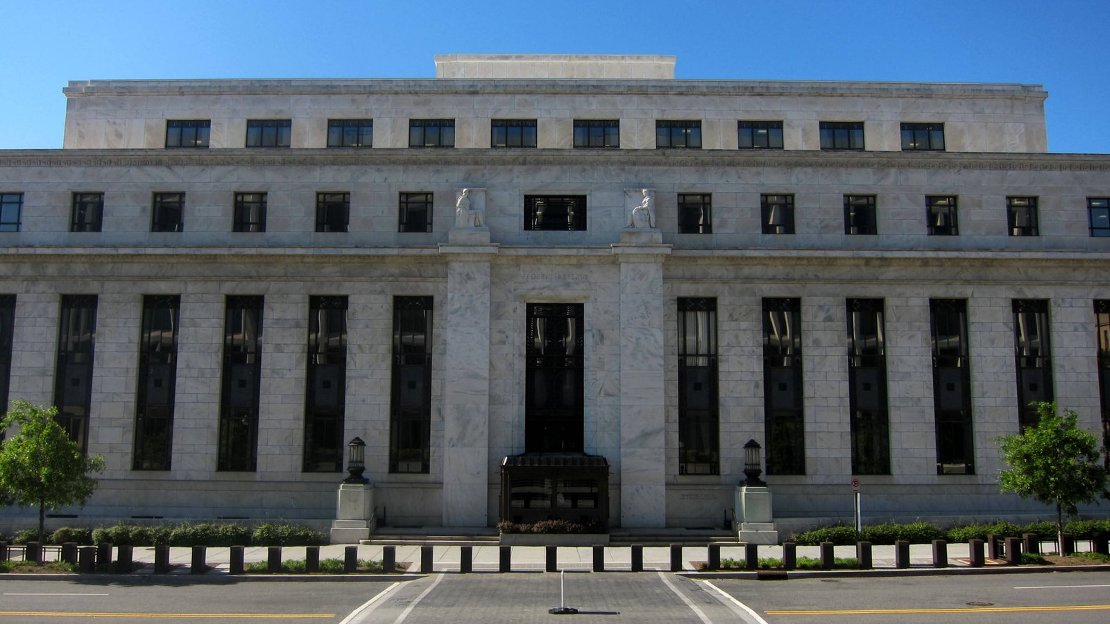The white marble north front of the Marriner S. Eccles Federal Reserve Board Building under a clear blue sky, with the words Federal Reserve carved above the central doorway and an empty street in front.