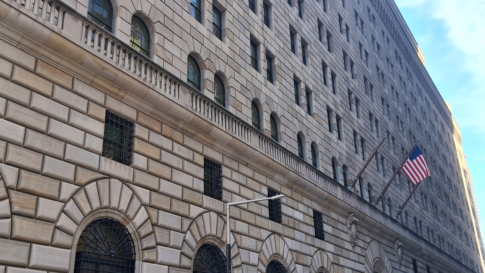 The stone facade of the Federal Reserve Bank of New York building in Manhattan, with arched windows and a United States flag.