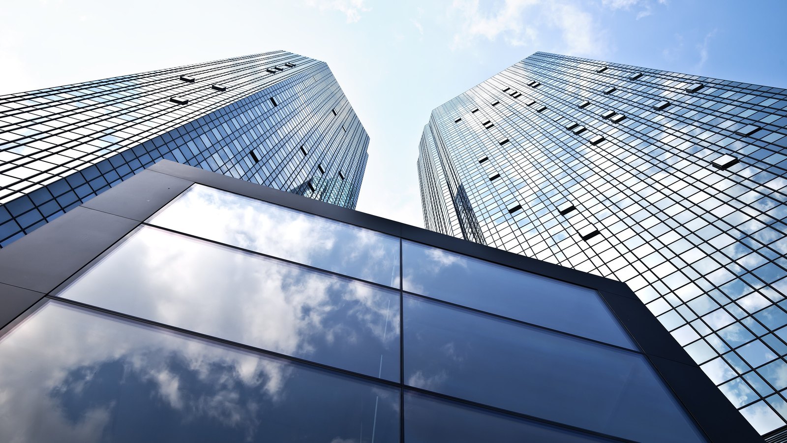 The two glass towers of the Deutsche Bank headquarters on the Taunusanlage in Frankfurt, photographed from ground level looking straight up, with a lower glass canopy in the foreground reflecting white cloud and blue sky. No people, lettering or signage is in the frame.