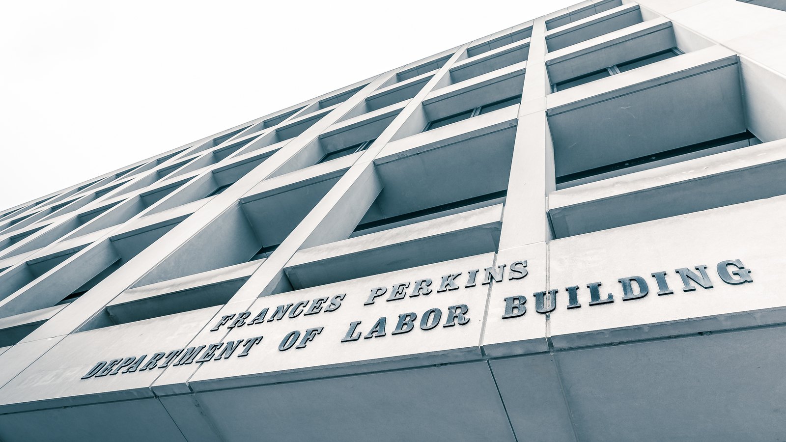 The corner of the Frances Perkins Building photographed steeply from below against a white sky, its grey piers and recessed window bays receding to a point. The inscription cut into the stone band across the facade reads FRANCES PERKINS DEPARTMENT OF LABOR BUILDING.