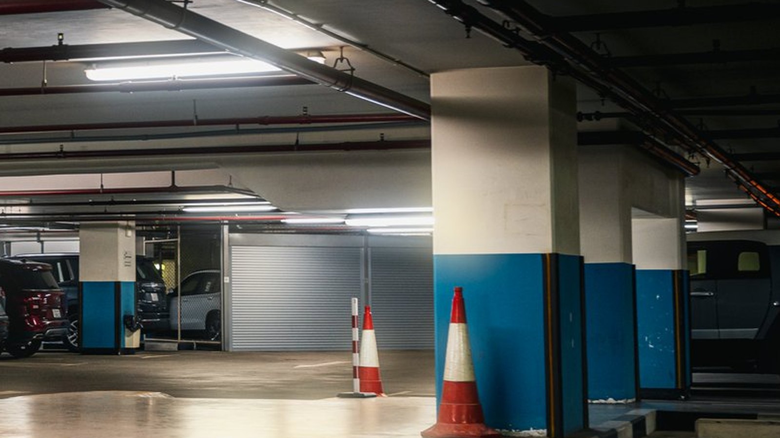 An underground parking garage under fluorescent light, with blue and white painted columns, a closed roller shutter and two red and white traffic cones; a few parked cars sit in the shadows with no legible plates, signs or markings. A generic parking illustration, not a photograph of this event.