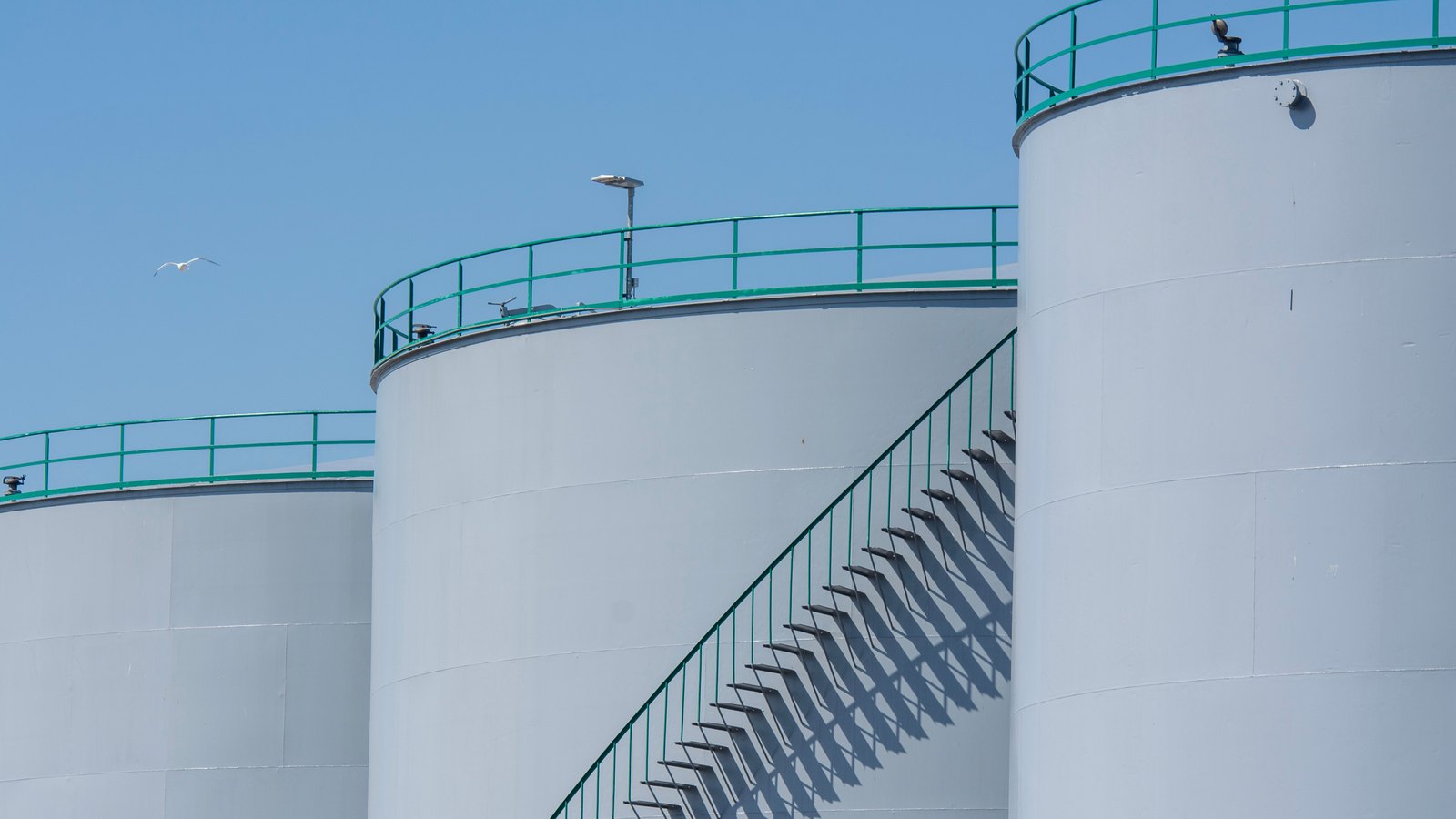 Three white cylindrical oil storage tanks with green handrails and an external stairway, seen against a clear blue sky.