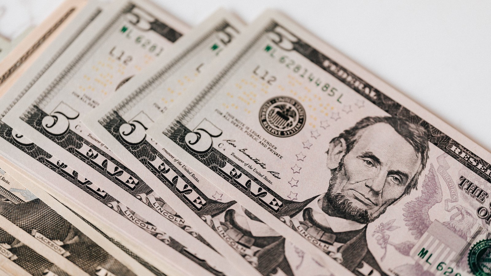 Overlapping United States five-dollar bills in a close crop, with Abraham Lincoln's portrait and Federal Reserve seals visible.