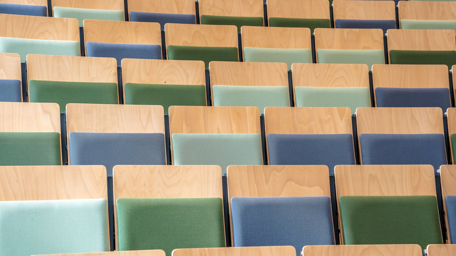 Rows of empty auditorium seats with pale wooden backs and green, mint and blue upholstered seats, repeating across the whole frame.