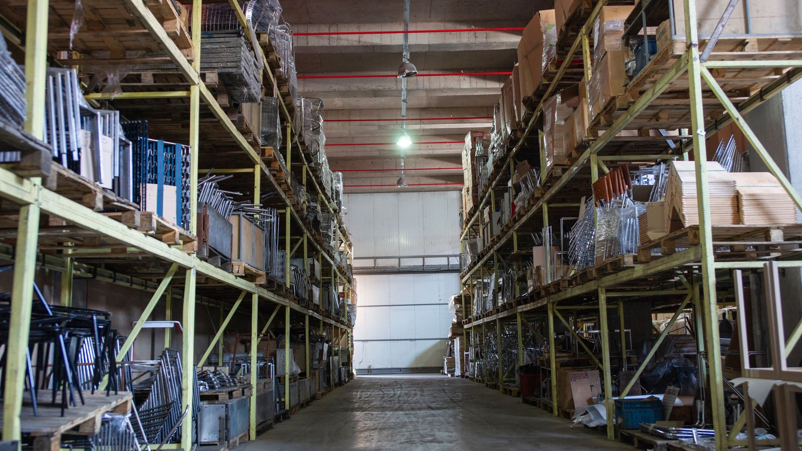 The central aisle of a warehouse lined with tall pallet racking holding boxes and metal goods.