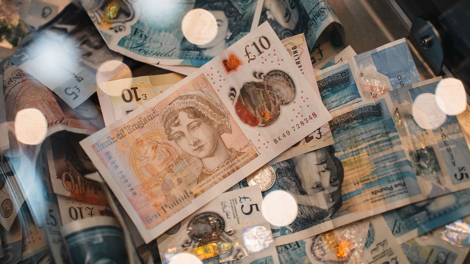 A loose scatter of Bank of England ten and five pound polymer notes with Royal Bank of Scotland fives beneath them, a coin resting on top.