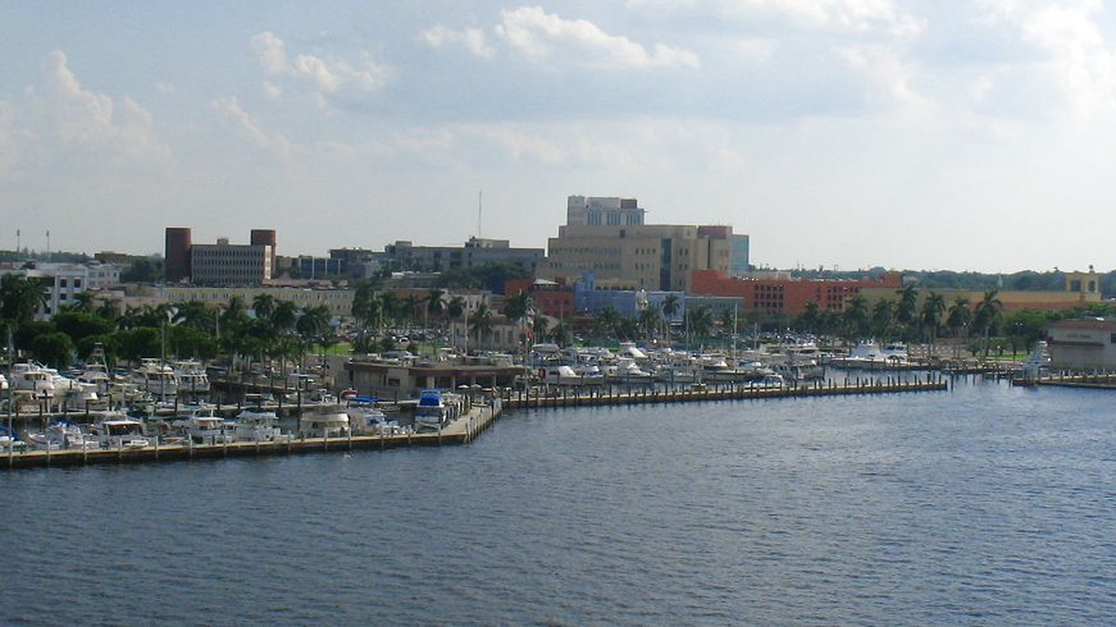 Downtown Fort Myers seen across the Caloosahatchee River. A long marina of moored white boats and wooden docks runs along the near bank behind a line of palm trees, with low and mid-rise buildings in tan, red and blue standing behind it. Open grey-blue water fills the lower half of the frame under a bright, partly clouded sky. No people, signage, text or place name is legible.