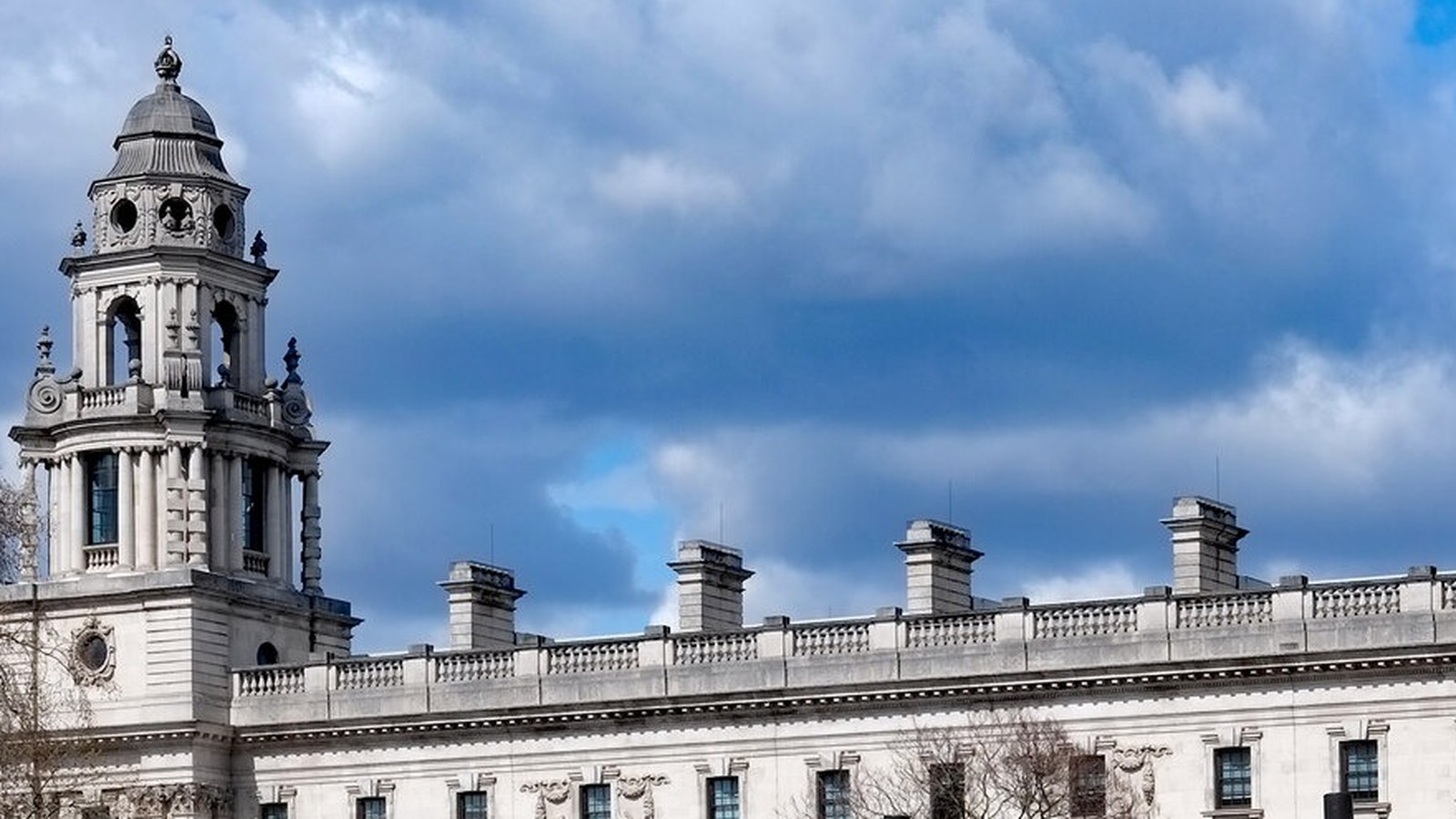 The domed corner tower and stone balustraded roofline of the Government Offices Great George Street building in Westminster, home to HM Treasury, under a blue sky with heavy grey cloud.