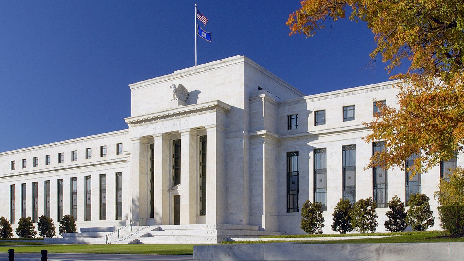 The Marriner S. Eccles Federal Reserve Board Building in Washington seen from an angle in autumn light, its white marble portico against a deep blue sky, with the United States flag flying above the roofline.