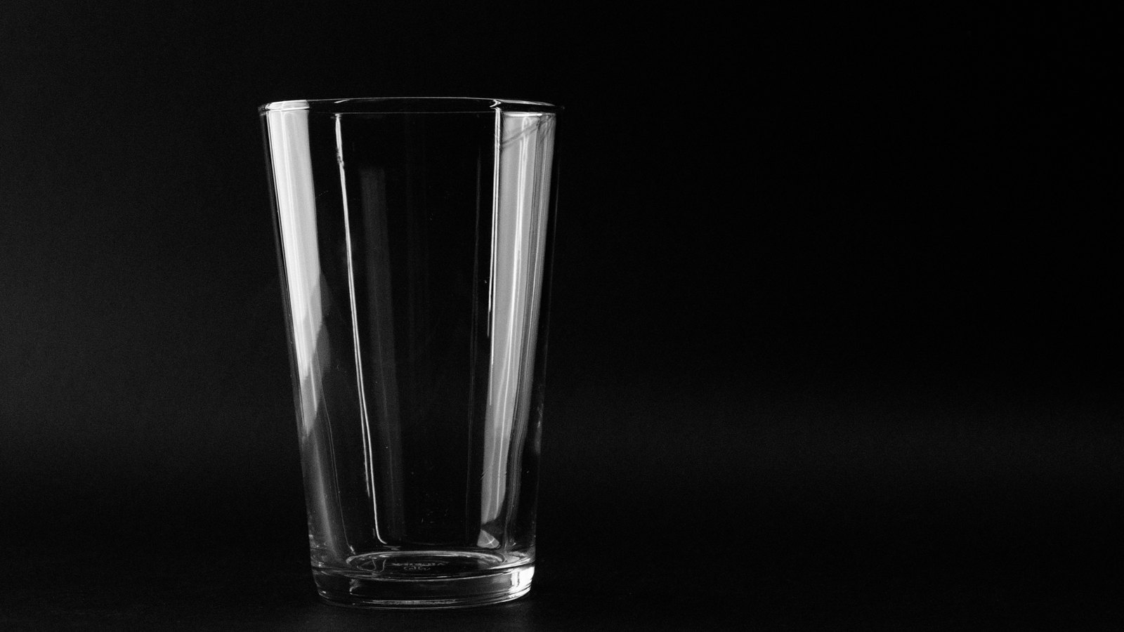 A black and white photograph of a single empty clear drinking glass standing on a dark surface against a black background, lit from the left so the rim and one side catch the light. The glass holds no water. There is a faint embossed maker's mark near its base which is not legible at any size, and no other text, signage or lettering appears anywhere in the frame.