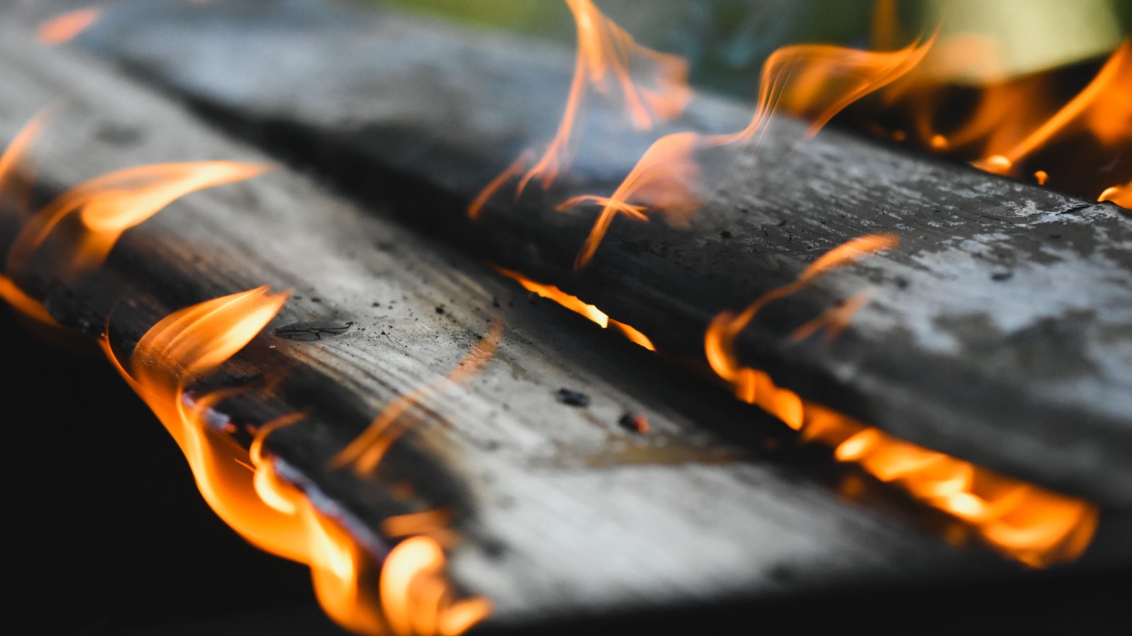 A close crop of two weathered grey timber planks with small orange flames running along their edges and blackened charring across the grain, shot against a dark blurred background, with no people, buildings, vehicles, signs or text in view. A generic illustration, not a photograph of this fire.