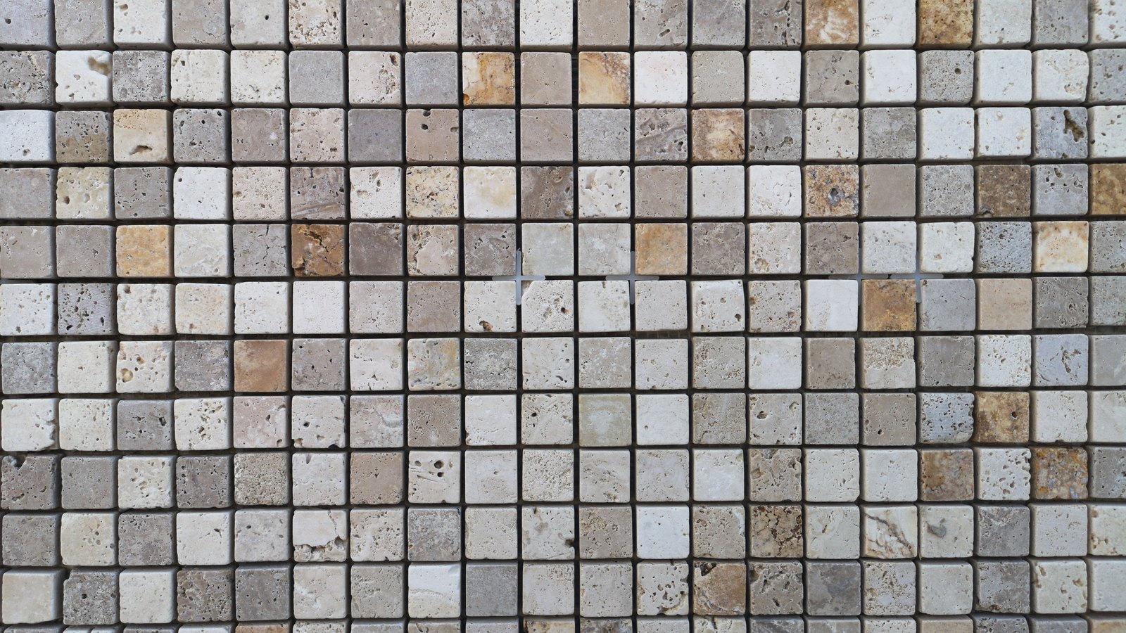 A dense grid of hundreds of small square travertine mosaic tiles filling the whole frame, in mixed cream, grey and pale brown tones with pitted natural stone surfaces and thin dark grout lines between them. No text, no logo, no person and no identifiable place appears in the image.