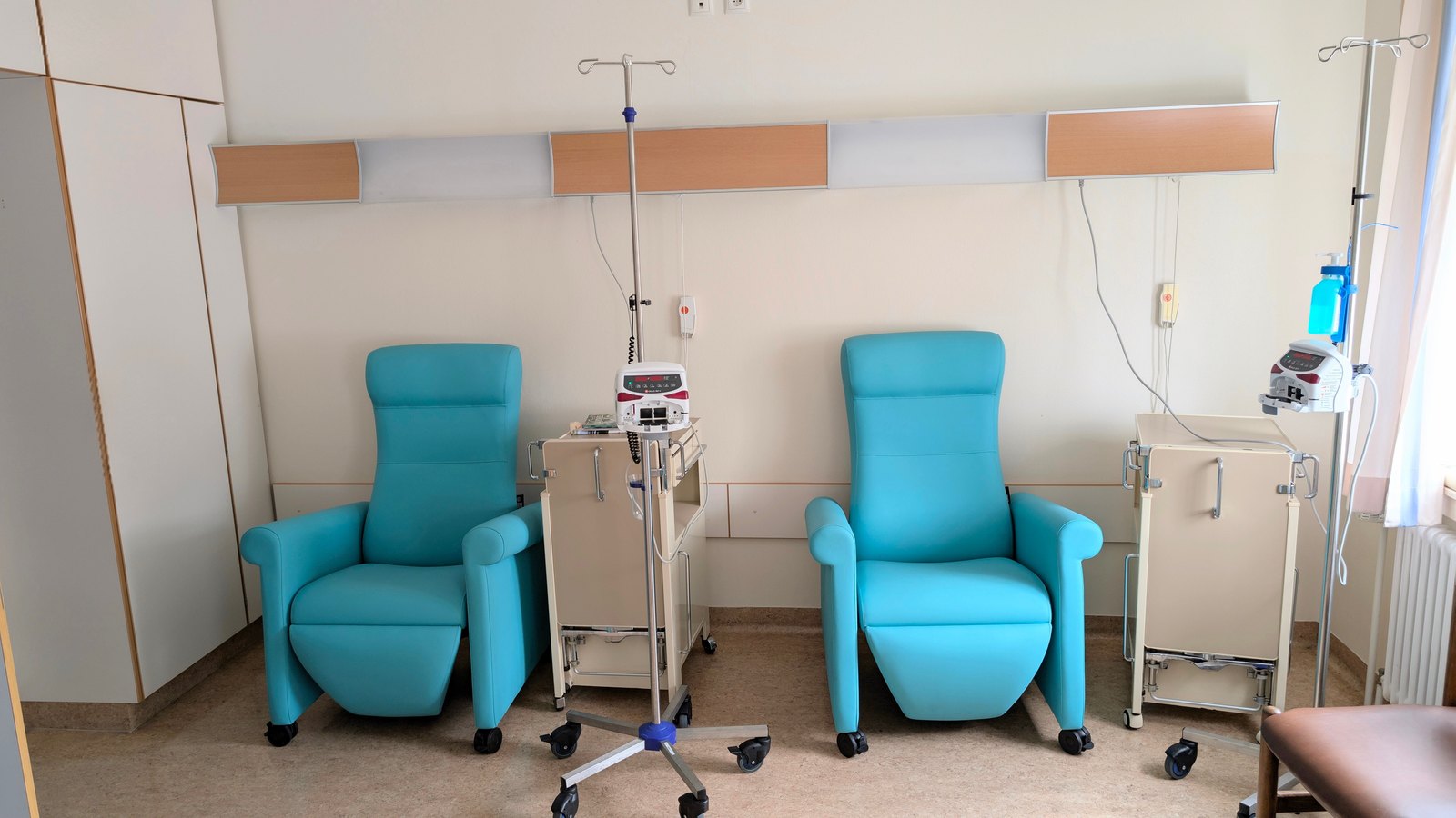 An empty treatment room with two turquoise reclining infusion chairs, IV poles and pumps against a pale wall, with no people or legible signs. A generic infusion room, not a photograph of any American Oncology Network clinic.