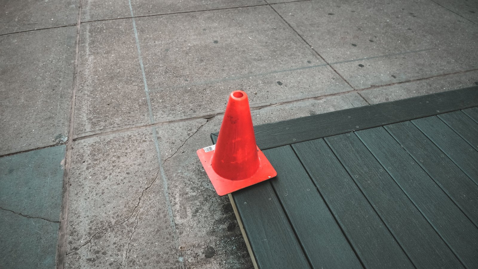 A single small orange traffic cone standing where a weathered, cracked concrete pavement meets a run of grey-green composite decking, seen from above in flat overcast light, with no people, vehicles, buildings, signs or text in view.