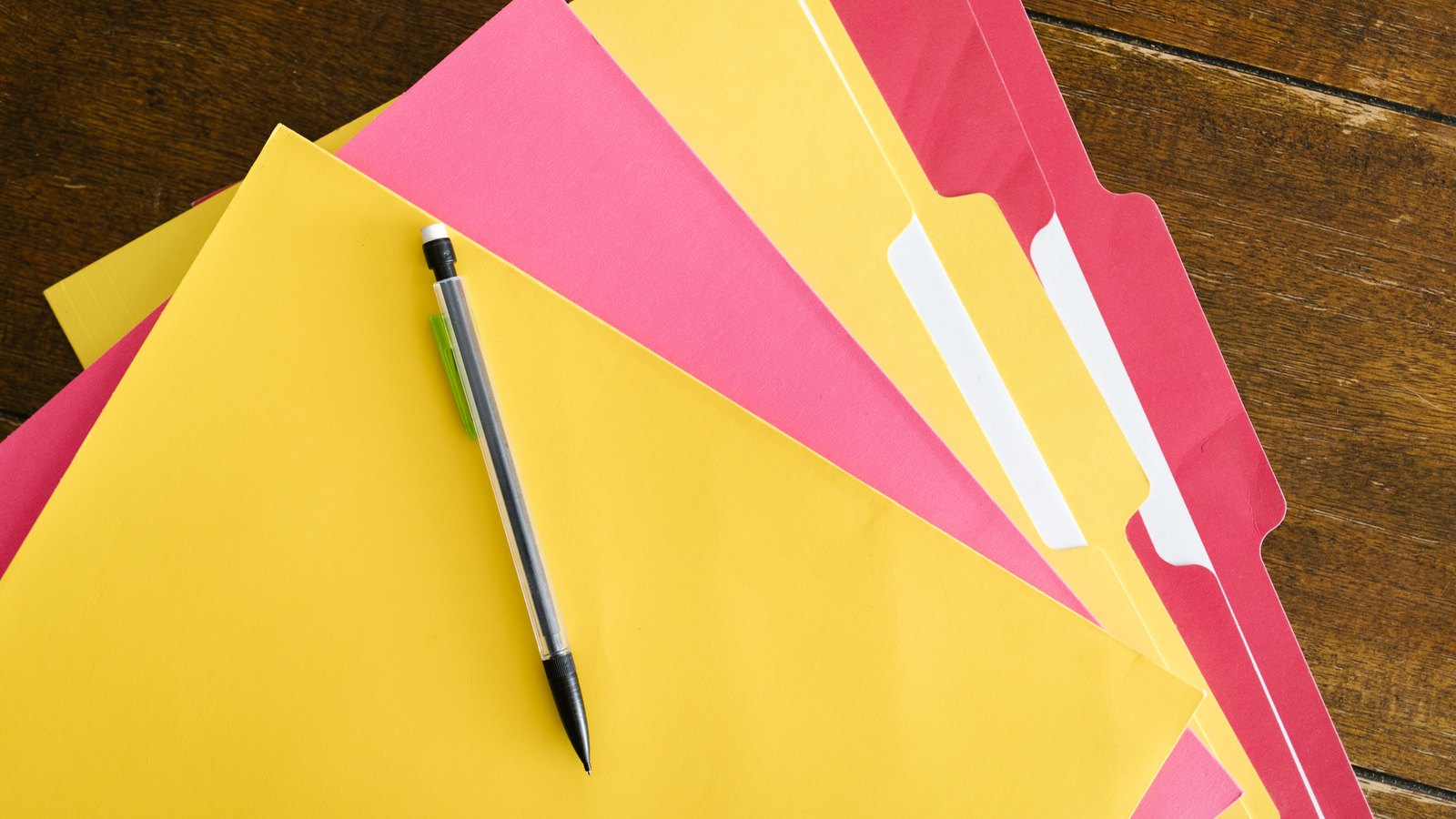 Five file folders in yellow and pink fanned out across a dark wooden table, with a clear-barrelled mechanical pencil resting on the topmost one. The folder tabs are blank and the frame carries no text, no logo, no signage and no person. It is a stock photograph of empty folders, not a photograph of any Fort Myers Beach record, office or person named in this item.