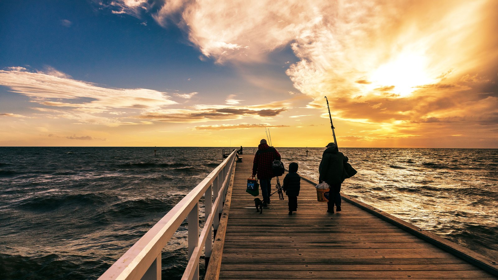 Three people seen from behind in silhouette walking away along the timber deck of a fishing pier at sunset, carrying rods and tackle bags, a white handrail running down the left side, open sea flat to the horizon on both sides and a broken orange and gold cloud bank above. Not a photograph of this event.