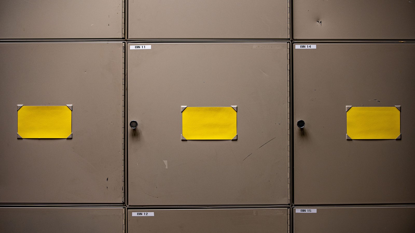 A wall of closed taupe metal storage cabinet doors photographed straight on. Three of the doors carry a blank yellow card held at its corners by metal clips, and two have a small round handle. Narrow white labels along the horizontal seams read BIN 11, BIN 12, BIN 14 and BIN 15. The metal is scuffed and lightly scratched. No person, no place name, no logo and no other text appears in the frame.