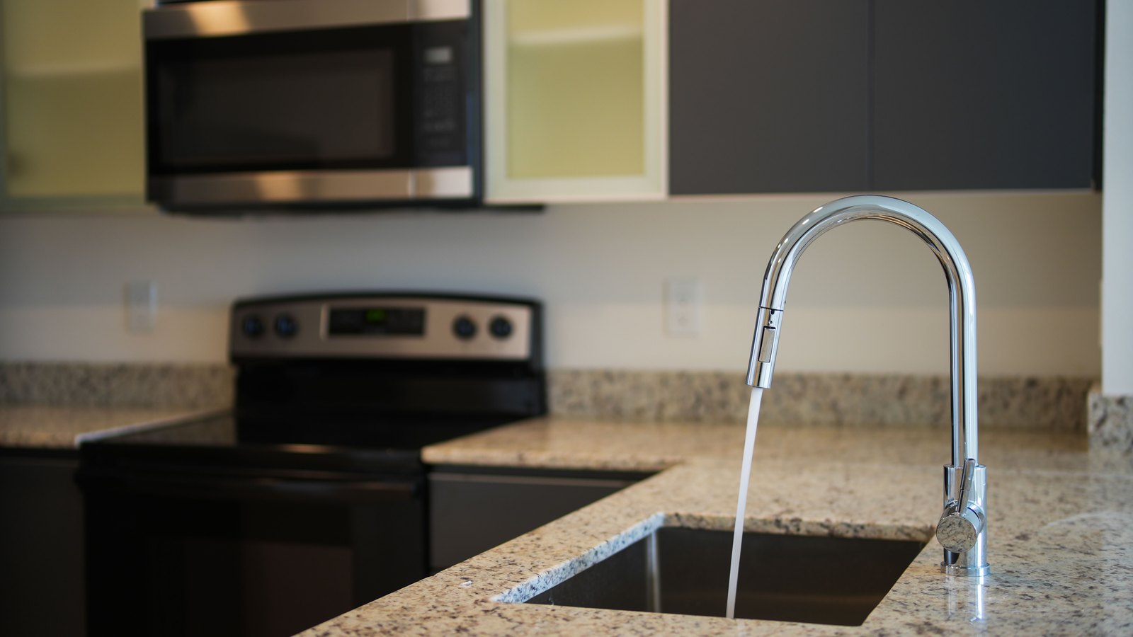 A chrome pull down kitchen tap running a steady stream of water into an undermount sink set in a speckled granite worktop, with a cooker and microwave out of focus behind it, and no people, signage or legible text in view. A generic illustration, not a photograph of Fort Myers.