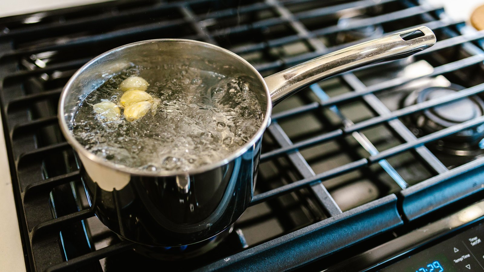 A small stainless steel saucepan of vigorously boiling water, with three garlic cloves floating at the surface, sitting on the black cast iron grate of a gas hob. The only legible text is the cooker's own control panel. A generic illustration, not a photograph of Fort Myers.