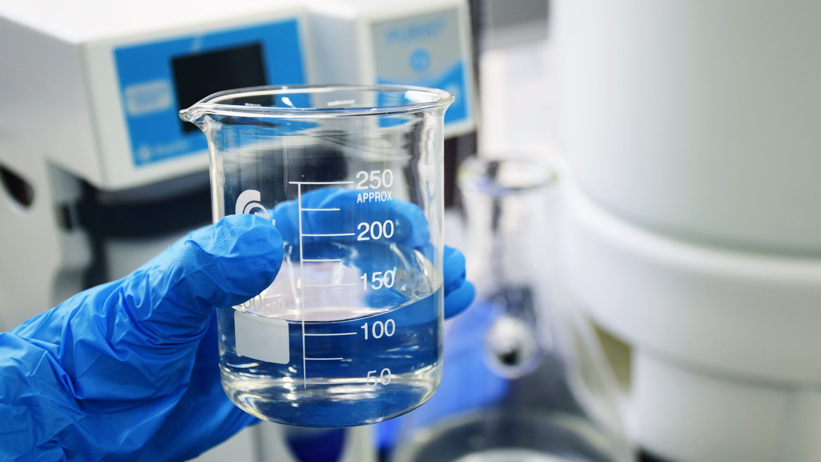 A hand in a blue nitrile glove holding a graduated 250 millilitre glass beaker part filled with a clear liquid, with laboratory instrument housings and a second piece of glassware out of focus behind it. No face, no name and no place signage is visible, and the only legible text is the volume scale moulded into the beaker. A generic illustration of laboratory testing, not a photograph of Fort Myers or of any Fort Myers water sample.