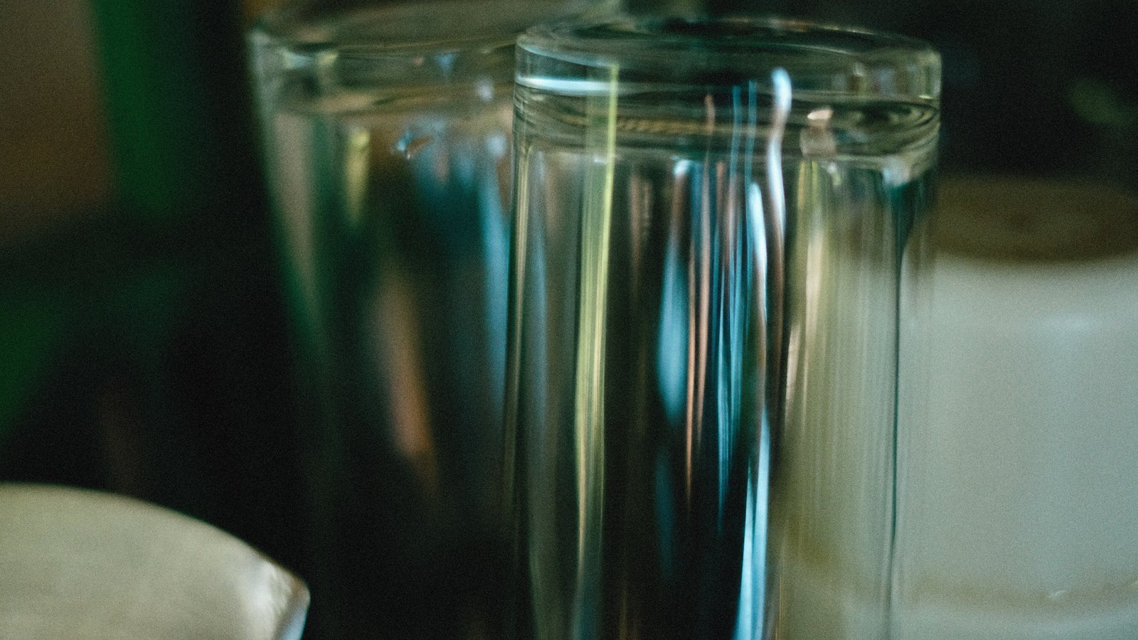 Two empty clear drinking glasses stand close together on a surface in dim domestic light, photographed close up, with no brand name, place name or legible text in view.