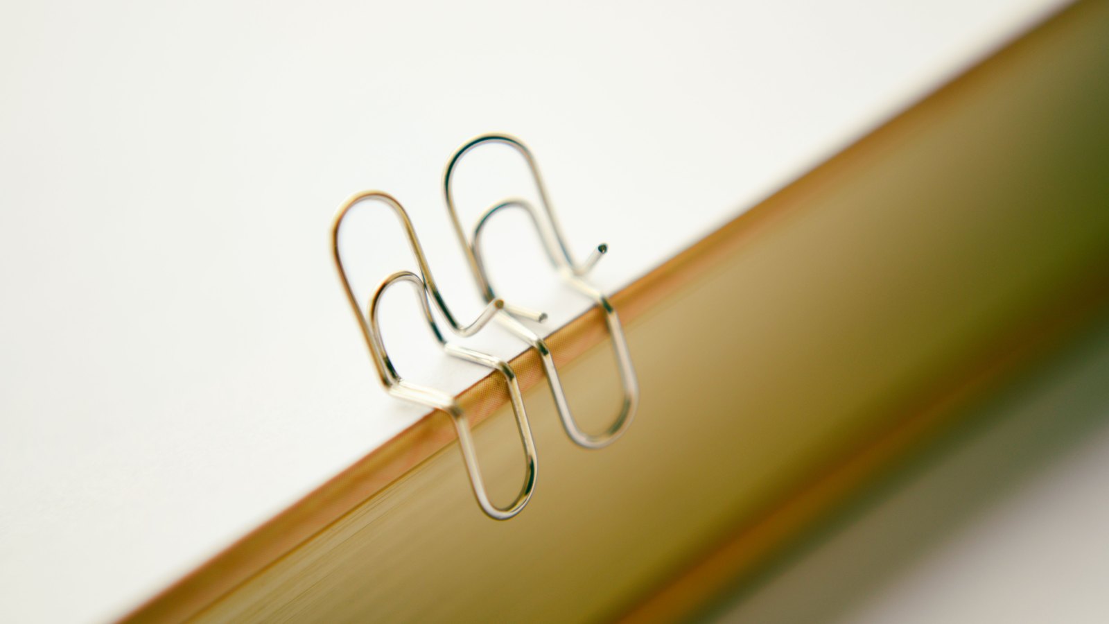 Two slim silver paperclips hooked over the gilded page edge of a closed book, photographed close up against a plain pale background. No text, no logo, no place and no person appears in the frame.