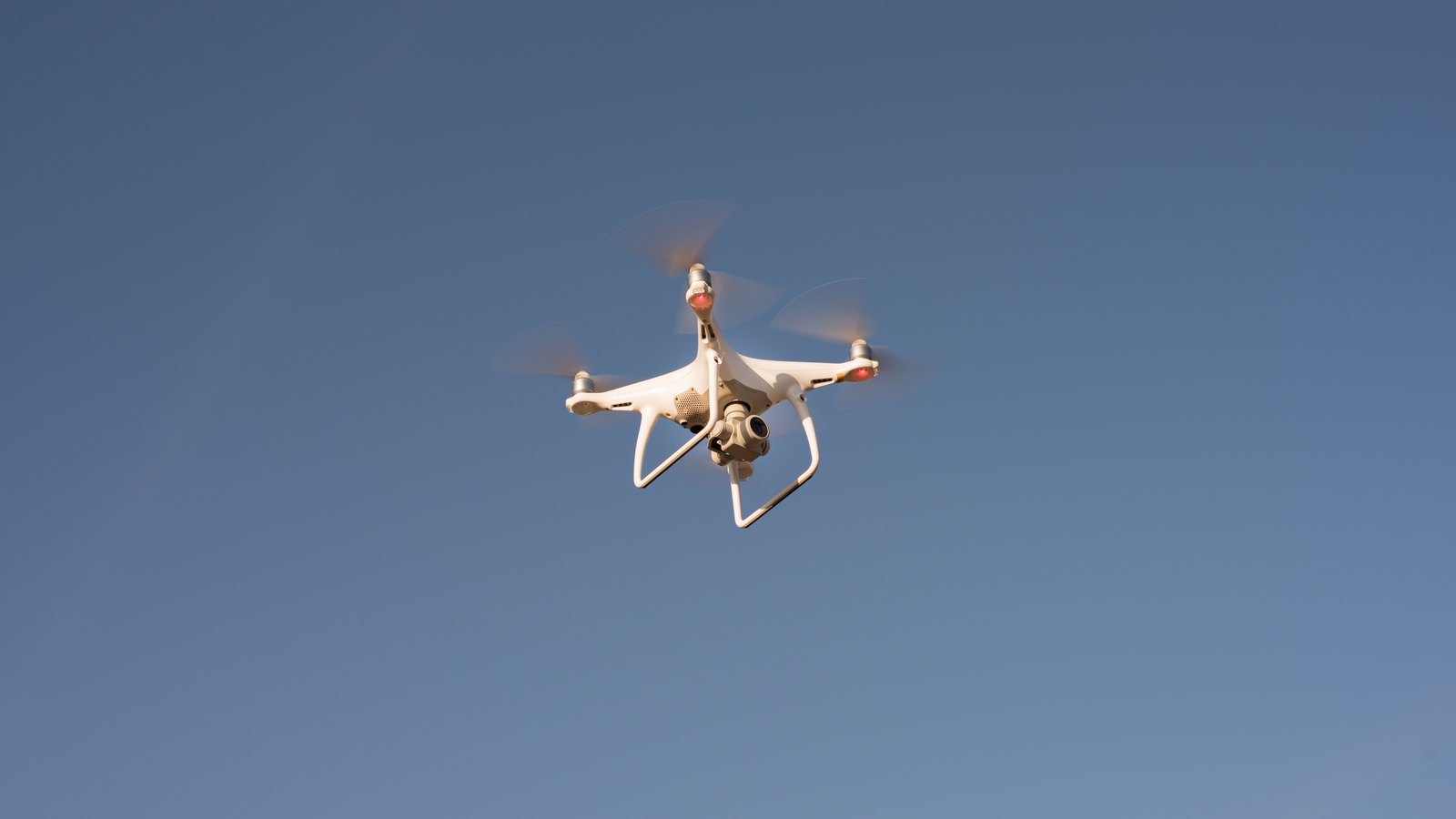A white four-rotor camera drone hovers against a clear blue sky, seen from below, with no people, buildings, markings or legible text in the frame. A generic drone illustration, not a photograph of this event.